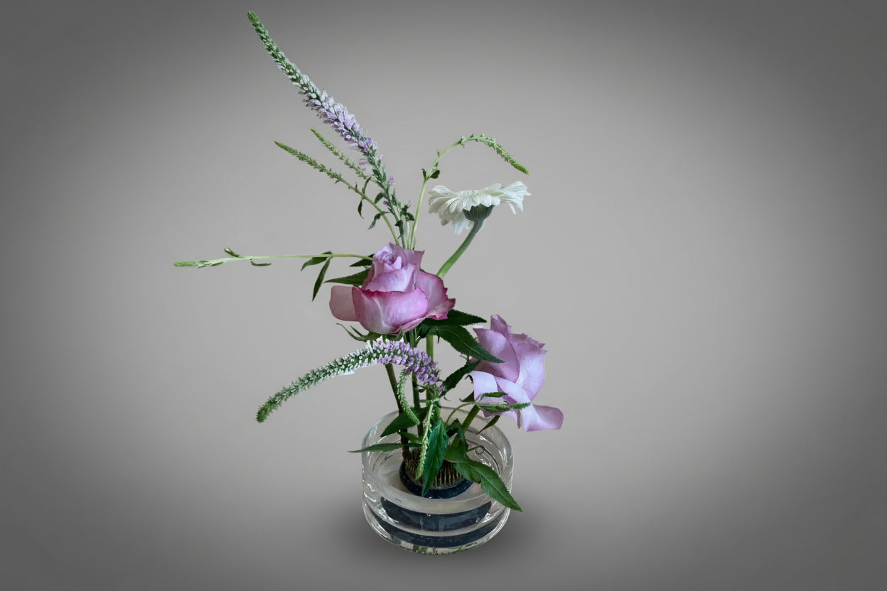 A floral arrangement featuring pink roses and other green and purple flowers in a custom shallow glass cylinder with a 'pin frog'.