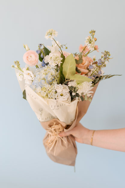 A medium vase and wrap arrangement of blue and white flowers, held by a person against a blue background.