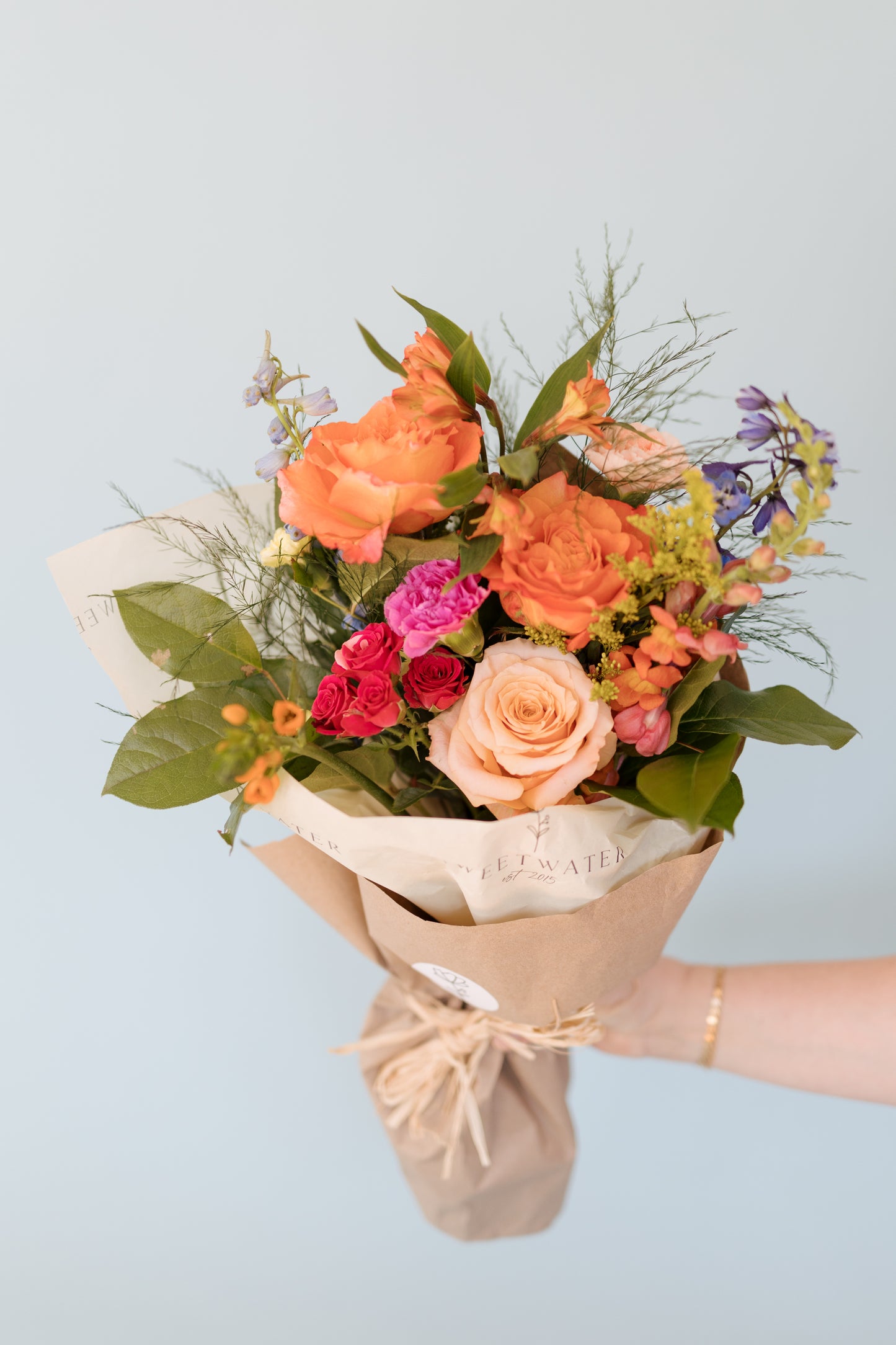 A bouquet of fresh flowers including shades of orange, pink, white, and green, wrapped in paper and twine.