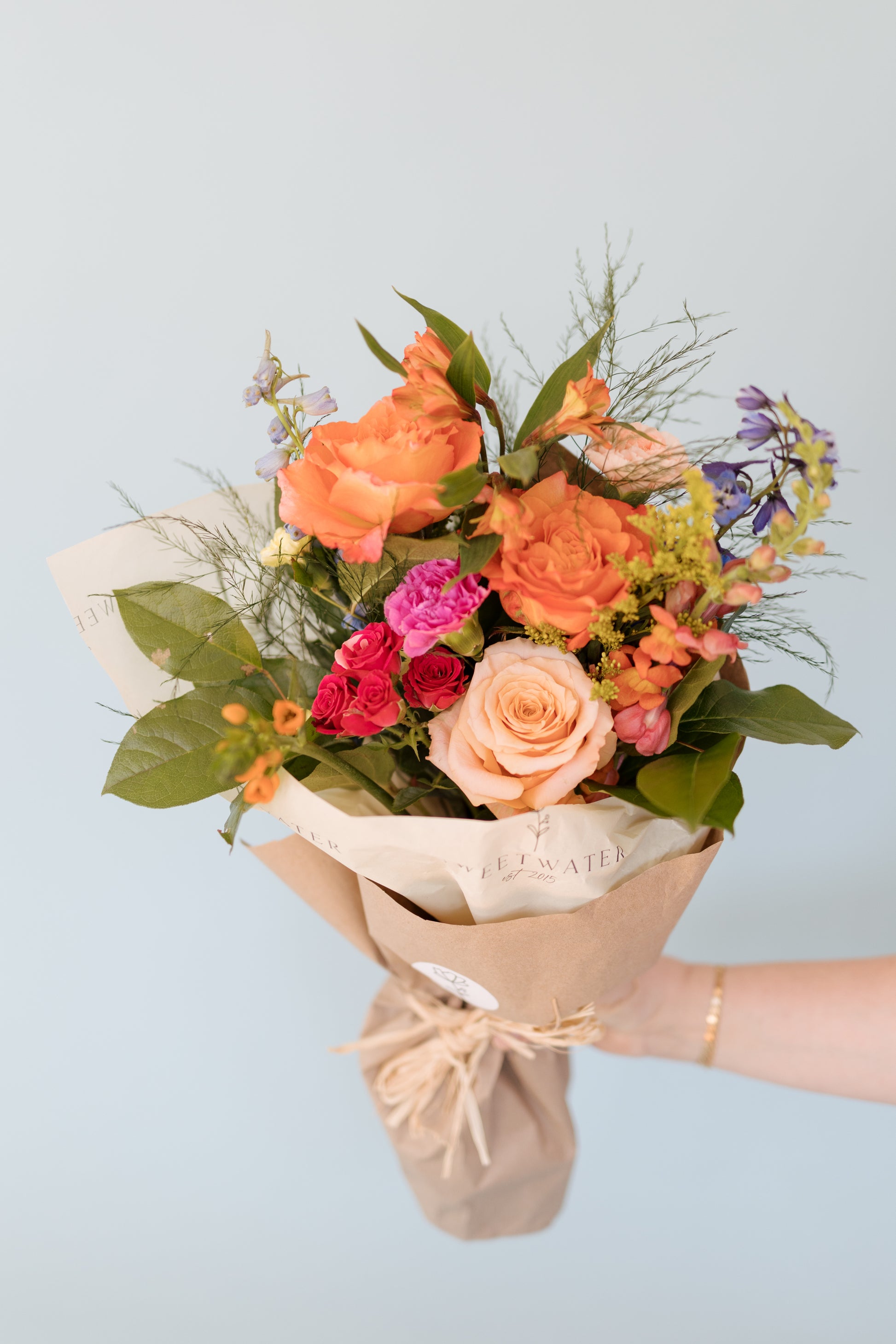 A bouquet of fresh flowers including shades of orange, pink, white, and green, wrapped in paper and twine.