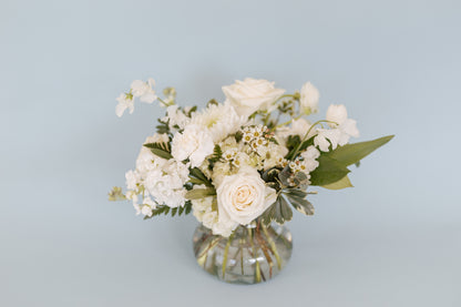 A medium-sized vase containing an assortment of white and green floral stems arranged in a classic style. The vase sits against a light blue background.