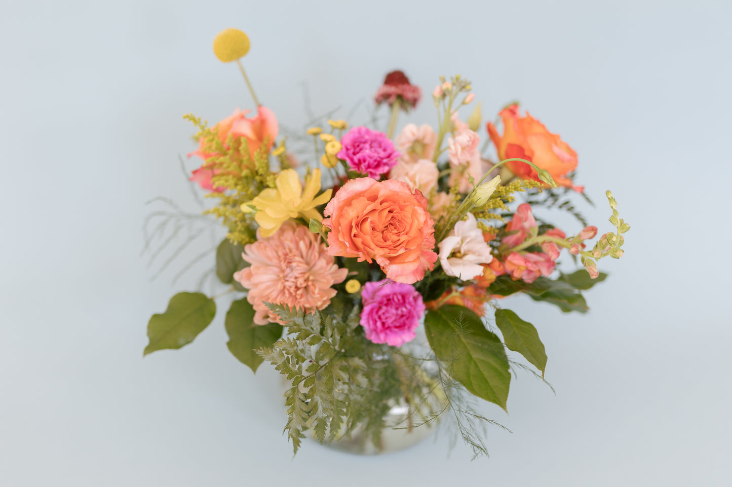 A colorful floral arrangement featuring a variety of flowers in shades of pink, peach, yellow, and green, presented in a clear round vase with a wrap bouquet.